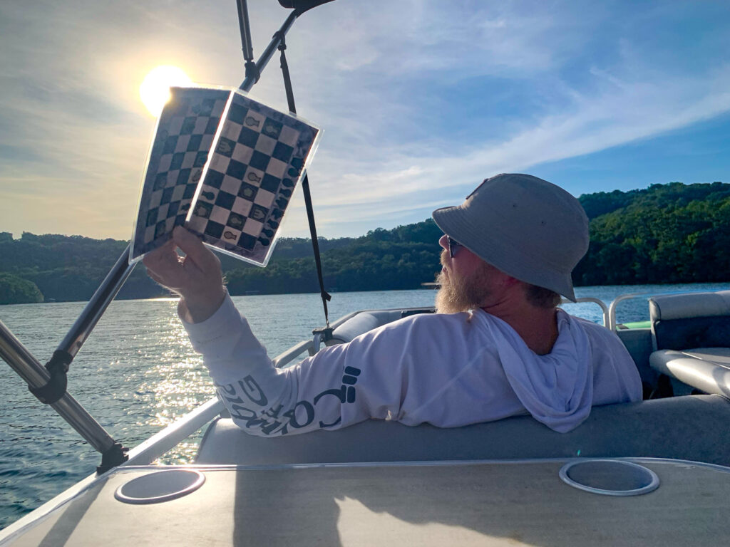 Man on a pontoon boat in the afternoon passing a travel chessboard back to the person taking the photo.