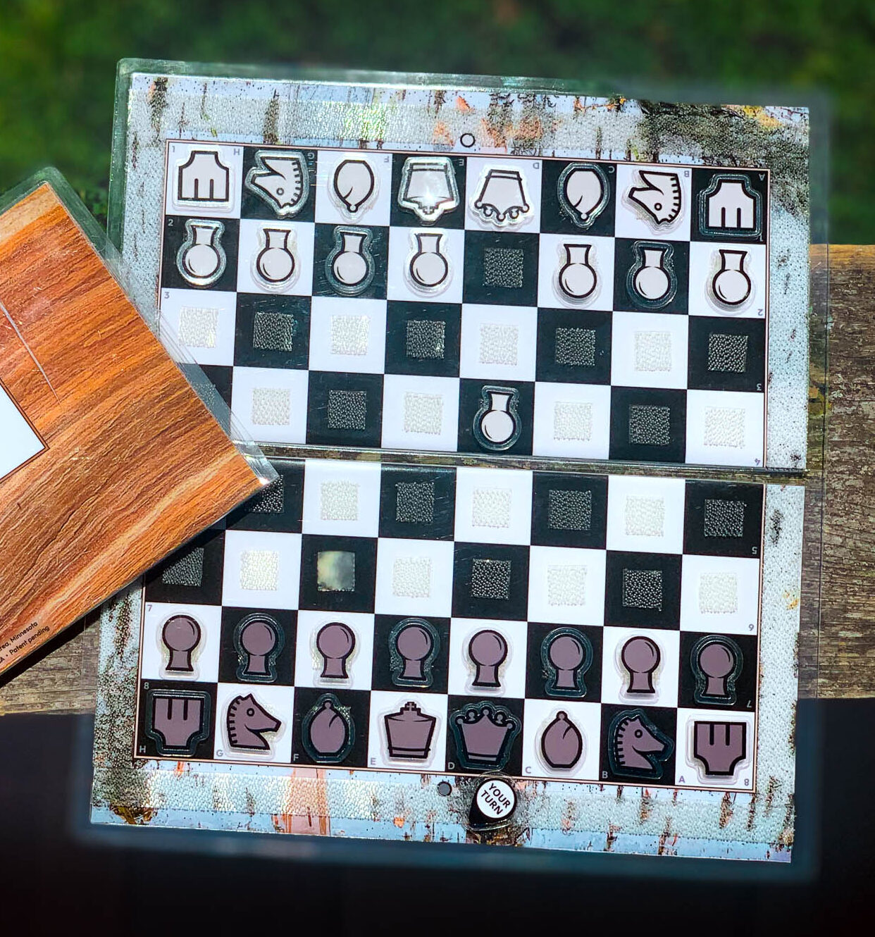 Close up of an open birch bark Travel Chessboard resting on a wooden rail in a park with a closed birch wood chessboard slightly out of the frame.