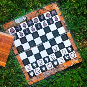 An open Travel Chessboard with Orange Lichen design with a game in progress sitting in grass. A closed Travel Chessboard with Wood design is slightly in the frame.