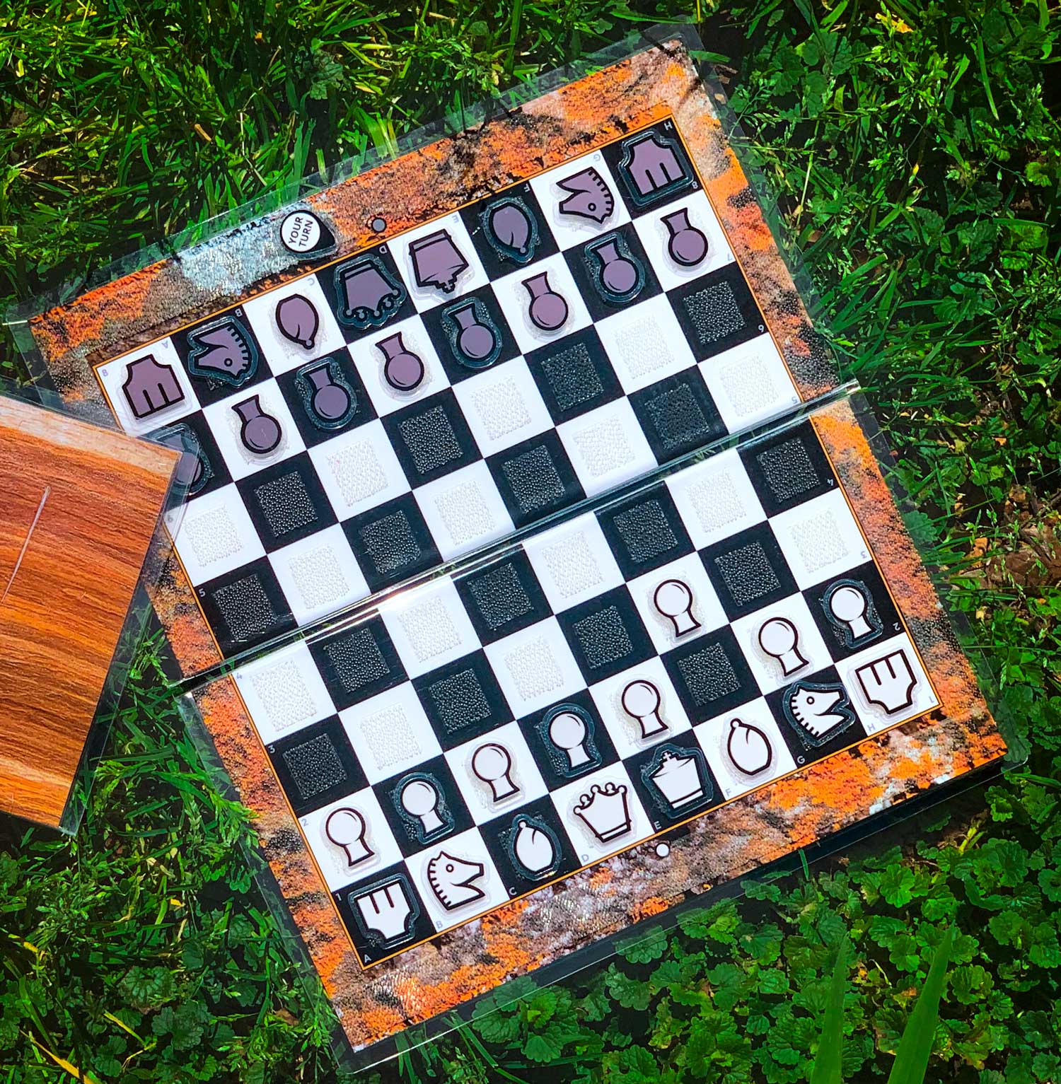 An open Travel Chessboard with Orange Lichen design with a game in progress sitting in grass. A closed Travel Chessboard with Wood design is slightly in the frame.