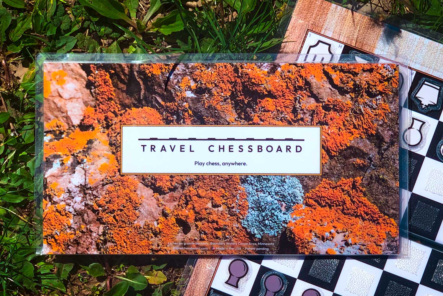 A closed Travel Chessboard with Orange Lichen design sitting half on grass and half on an open Travel Chessboard with Wood design.