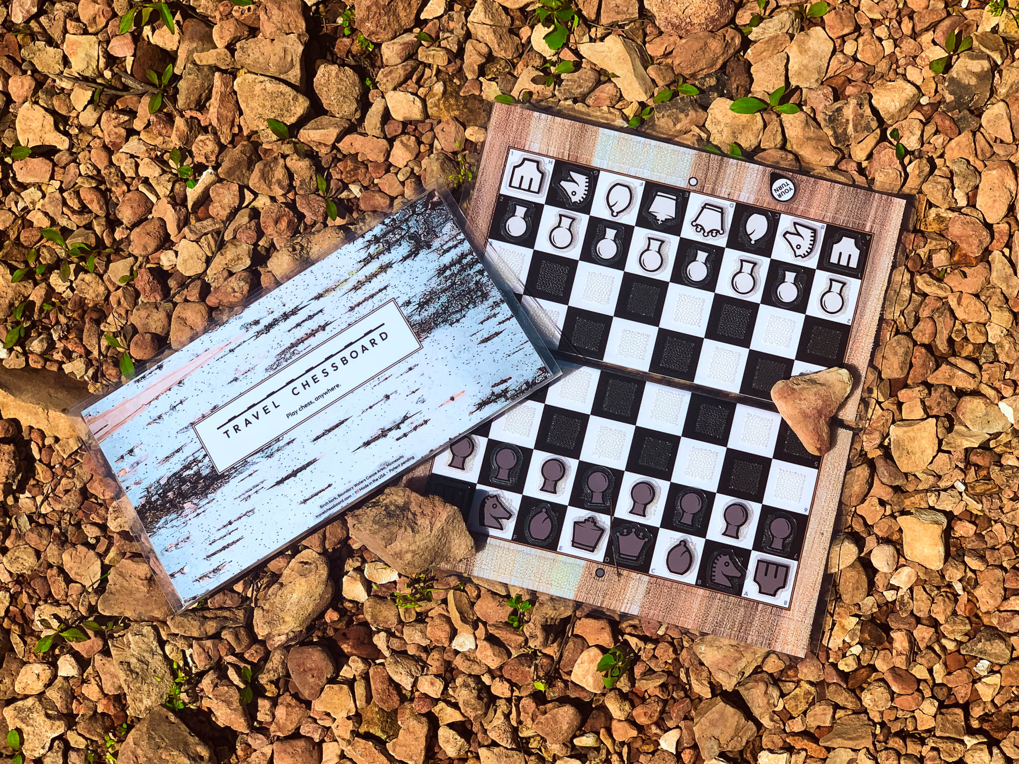 Open birch wood travel chessboard with closed birch bark chessboard on top and to the side, partially out of the frame, both on a bed of tan rocks.