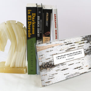 Closed Travel Chessboard with birch bark, leaning against a few books buttressed by a marble knight bookend, all on a white background.