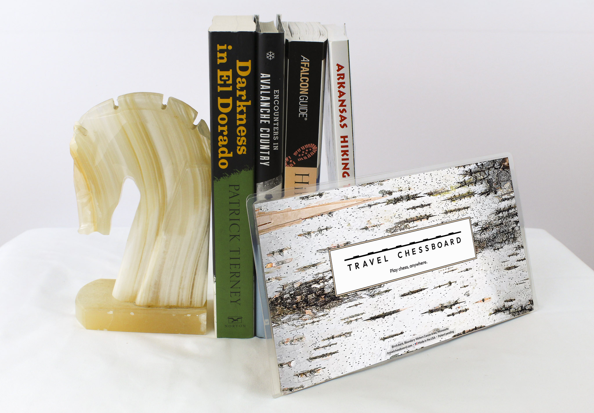 Closed Travel Chessboard with birch bark, leaning against a few books buttressed by a marble knight bookend, all on a white background.