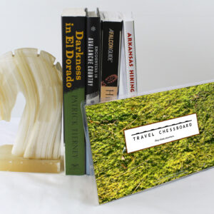 Closed Travel Chessboard with green moss, leaning against a few books buttressed by a marble knight bookend, all on a white background.