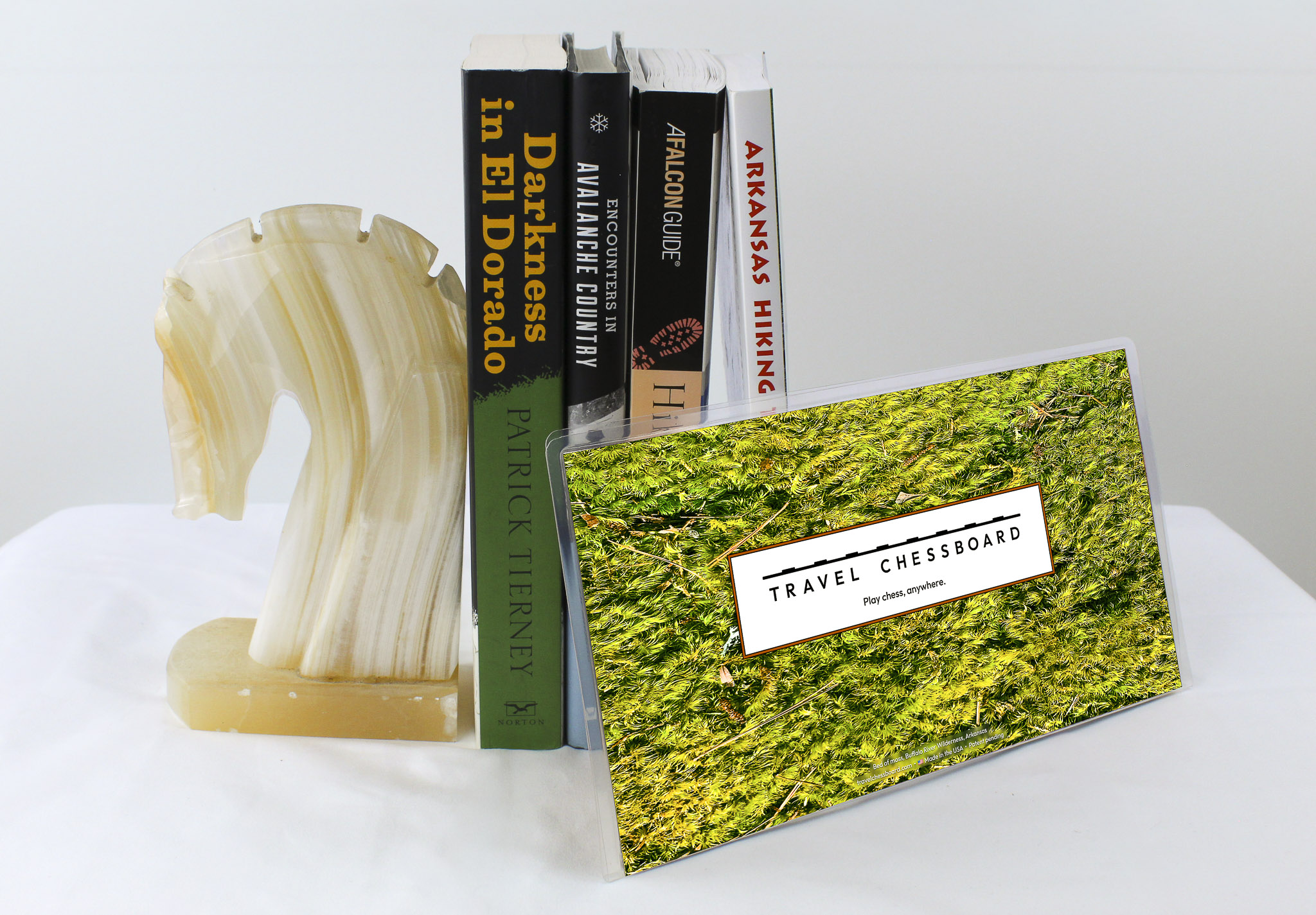 Closed Travel Chessboard with green moss, leaning against a few books buttressed by a marble knight bookend, all on a white background.