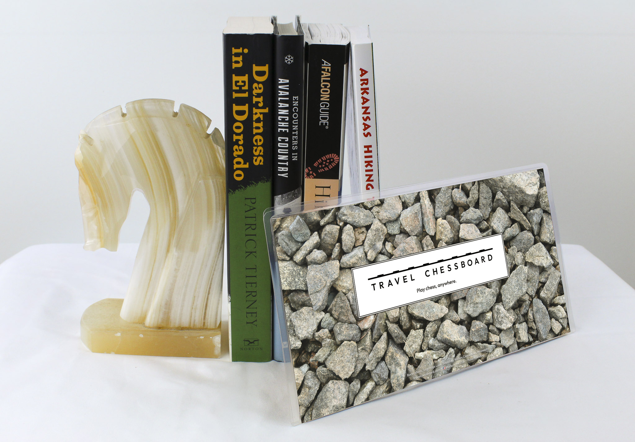 Closed Travel Chessboard with rocks, leaning against a few books buttressed by a marble knight bookend, all on a white background.