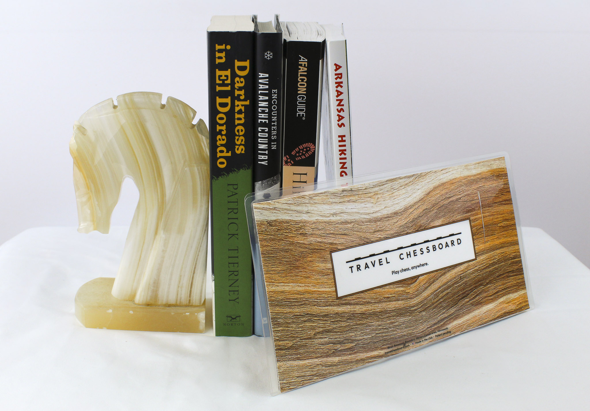 Closed Travel Chessboard with birch wood, leaning against a few books buttressed by a marble knight bookend, all on a white background.